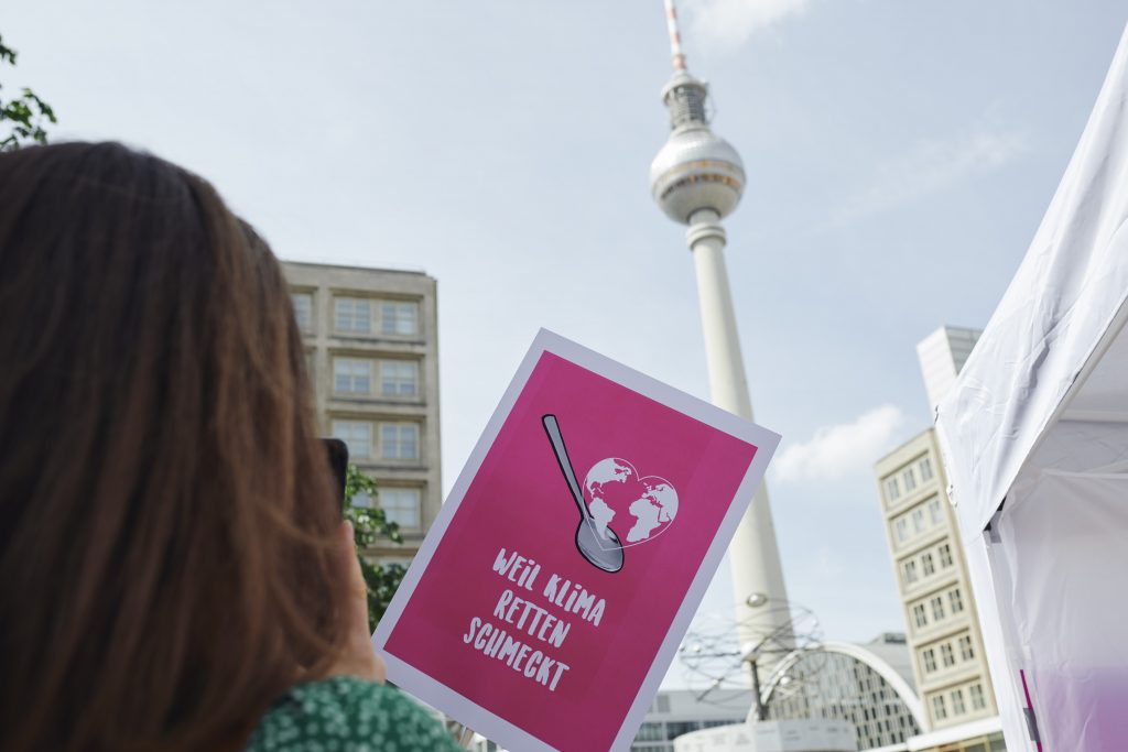 Auf einem Schild steht "Weil Klima retten schmeckt". Darüber ist als Illustration eine Erdkugel als Herz zu sehen und ein Löffel. Im Hintergrund sieht man den Fernsehturm in Berlin.