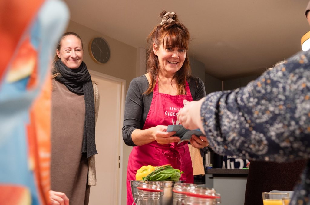 Eine Frau übergibt einer anderen etwas, eingewickelt in eine Serviette. Sie nimmt es lächelnd entgegen. Eine weitere Frau im Hintergrund freut sich mit ihr.