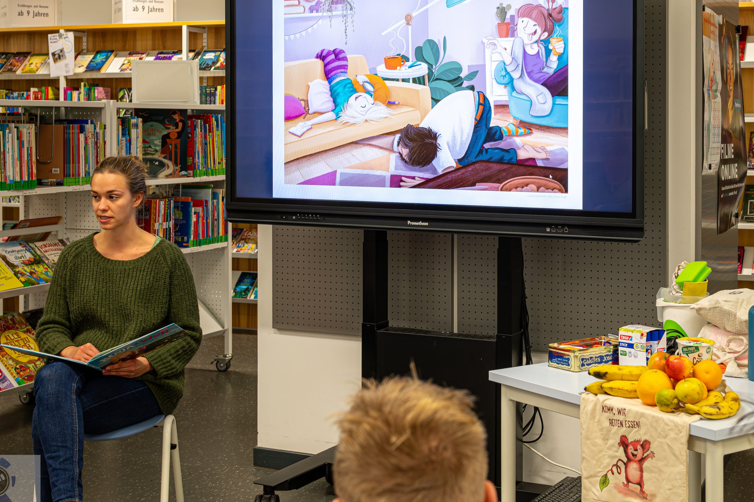 Eine junge Frau sitzt vor einer Gruppe von Kleinkindern und liest aus dem Kinderbuch "Benja und Wuse - Essensretter auf großer Mission" vor. Hinter ihr werden auf einem Bildschirm Bild-Ausschnitte aus dem Buch abgespielt.