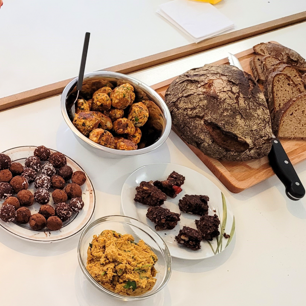 Auf einem Tisch stehen frisch geschnittenes Brot und verschiedene selbstgemachte Snacks: Linsentaler, cremiger Linsenaufstrich, schokoladige Energyballs und Schokoschnitten.