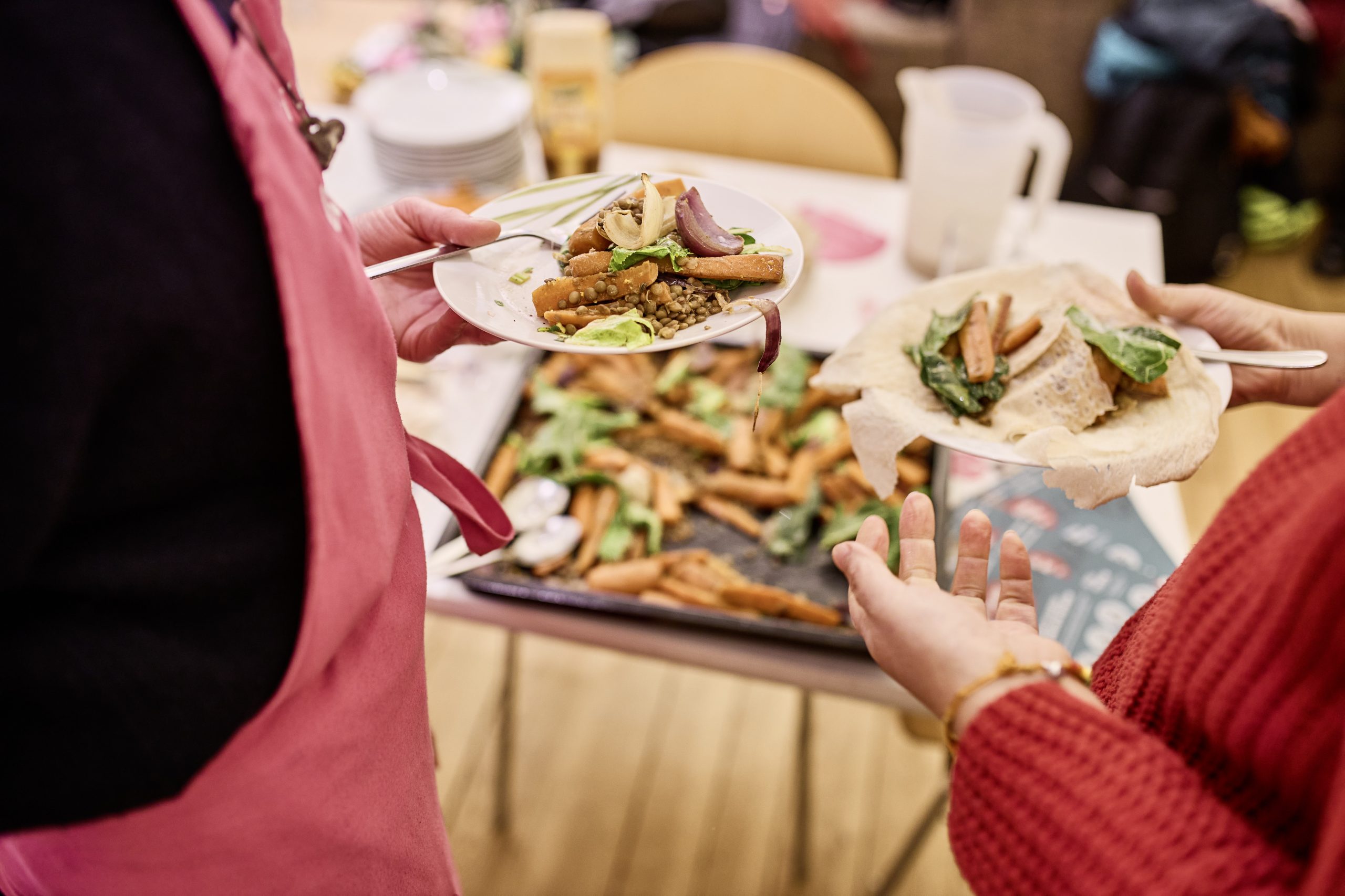 Zwei Personen halten je einen Teller mit Mittagessen in der Hand und unterhalten sich dabei. Im Hintergrund sind weitere Speisen zu sehen.