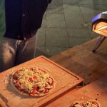 Ein Pizza-Ofen steht im Freihen auf einem Tisch. Davor sind zwei fertige Pizzen auf dem Tisch.