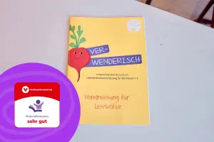 Die VERWENDERISCH Handreichung liegt auf einem Tisch. Auf dem Foto ist das Auszeichnungslogo des Materialkompass mit Note "sehr gut" zu sehen.