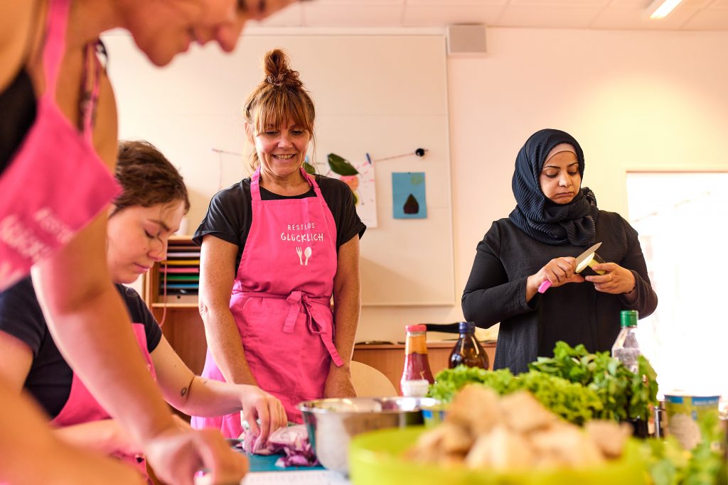 4 Frauen kochen zusammen - 3 tragen pinke schürzen mit dem Logo von RESTLOS GLÜCKLICH, 1 trägt ein Kopftuch. Im Vordergrund sind unscharf Lebensmittel zu sehen.