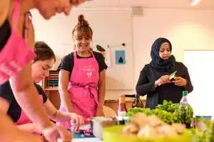 4 Frauen kochen zusammen - 3 tragen pinke schürzen mit dem Logo von RESTLOS GLÜCKLICH, 1 trägt ein Kopftuch. Im Vordergrund sind unscharf Lebensmittel zu sehen.