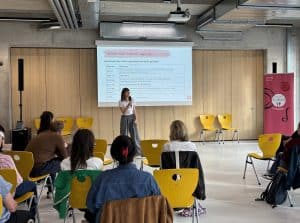 Mitarbeitende von RESTLOS GLÜCKLICH steht vor einer Gruppe von sitzenden Menschen und präsentiert spricht in eine Mikrofon
