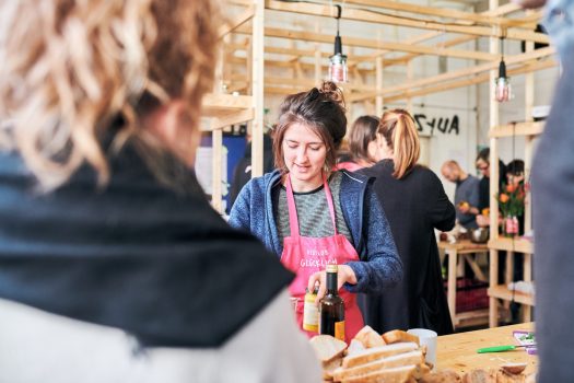 Ann-Kathrin Ridder von RESTLOS GLÜCKLICH e. V. stellt ein Glas Senf auf einem Tisch in einer Küche ab. Um sie herum und im Hintergrund sind mehrere Teilnehmende des Koch-Workshops, die sich unterhalten und Lebensmittel schneiden.