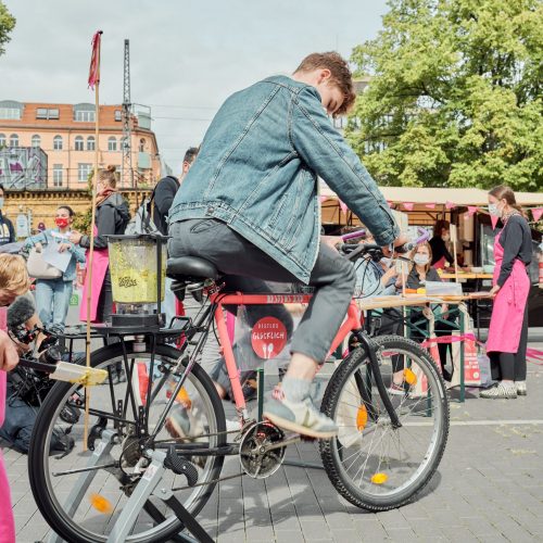 Die Gropiusstadt feiert Familiennacht mit vielen kostenlosen Mitmach-Aktionen – seid dabei! Bei einem Aktionsstand im Freien: Eine jugendliche Person sitzt auf dem Smoothiebike und tritt in die Pedale. Im Hintergrund ist der belebte Aktionsstand zu sehen.