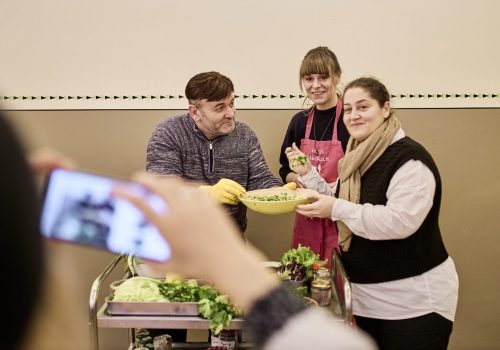 Eine Person hält ein Handy vor sich und fotografiert zwei Frauen und einen Mann, die nebeneinander stehen. Sie lächeln breit und zeigen die Gerichte, die sie im kostenlosen Koch-Workshop zubereitet haben, stolz in die Kamera.