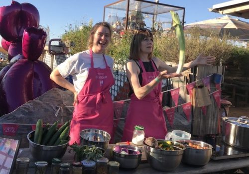 Zwei Frauen stehen nebeneinander und tragen pinke Kochschürzen. Eine von ihnen lächelt direkt in die Kamera. Die andere hält eine Stange Porree in der Hand und jongliert sie spielerisch.