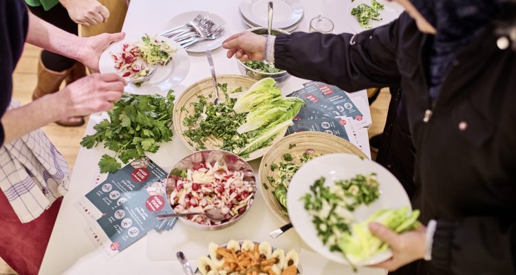 Auf einem Tisch stehen mehrere Schüsseln mit selbstgemachten Salaten, warmen Speisen und Beilagen. Menschen greifen zu und füllen ihre Teller.