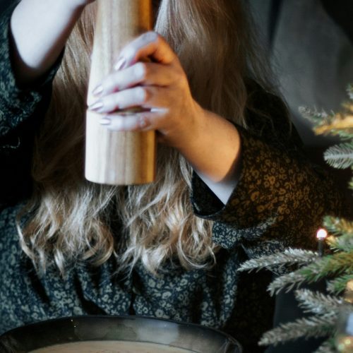 micheile-henderson-2ovx_PFxBiw-unsplash_bearbeitet Die Hände einer Person halten eine Pfeffermühle über einen Teller und malen Pfeffer. Rechts am Bildrand ist ein Weihnachtsbaum zu sehen.